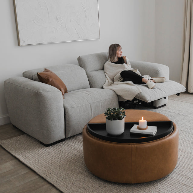 canape relax gris clair avec assise etendue, coussin brun et ambiance cocooning pres d une table ronde en cuir.