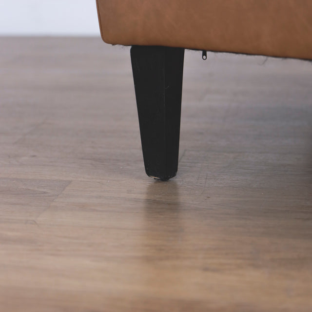 Image rapprochée d'une patte d'un fauteuil moderne en cuir végane cognac avec pieds noirs sur sol en bois.