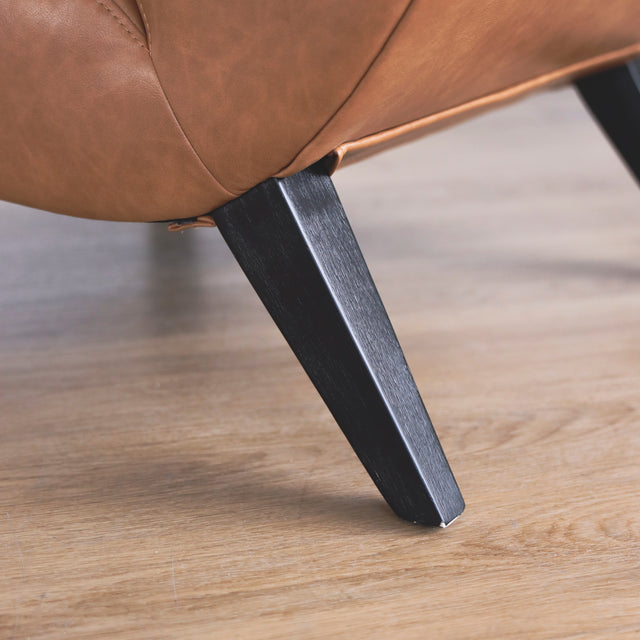 Image rapprochée d'une patte d'un fauteuil moderne en cuir végane cognac avec pieds noirs sur sol en bois.