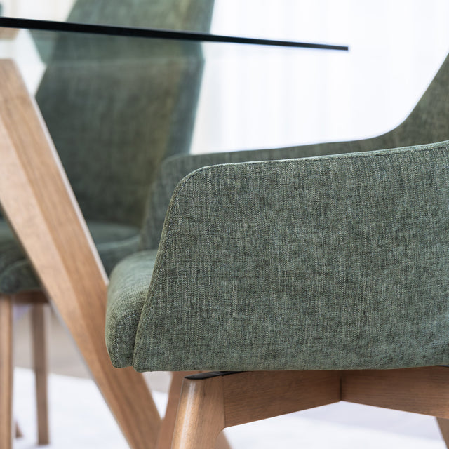 Chaise de salle à manger en tissu vert avec accoudoirs et pieds en bois, style moderne autour d’une table en verre.