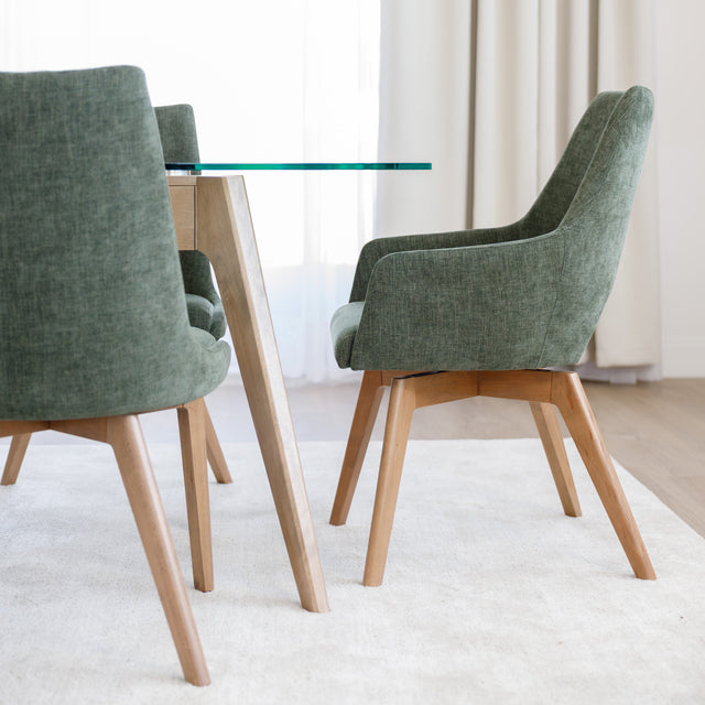 Chaise de salle à manger en tissu vert avec accoudoirs et pieds en bois, style moderne autour d’une table en verre.