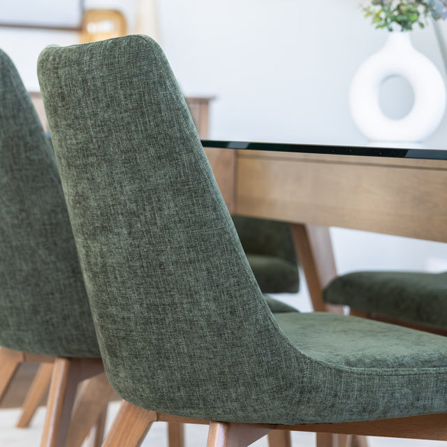 Chaise rembourrée en tissu vert avec dossier incurvé et pieds en bois, autour d’une table à manger au design moderne.