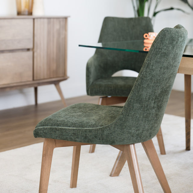 Chaise rembourrée en tissu vert avec dossier incurvé et pieds en bois, autour d’une table à manger au design moderne.