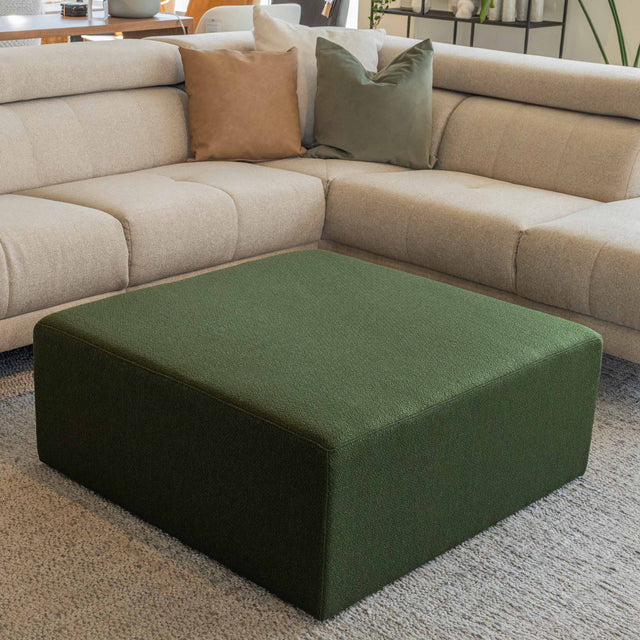 Ottoman carré vert en tissu, utilisé comme table basse devant un canapé d’angle moderne dans un salon contemporain.