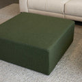 Ottoman carré vert en tissu, utilisé comme table basse devant un canapé d’angle moderne dans un salon contemporain.
