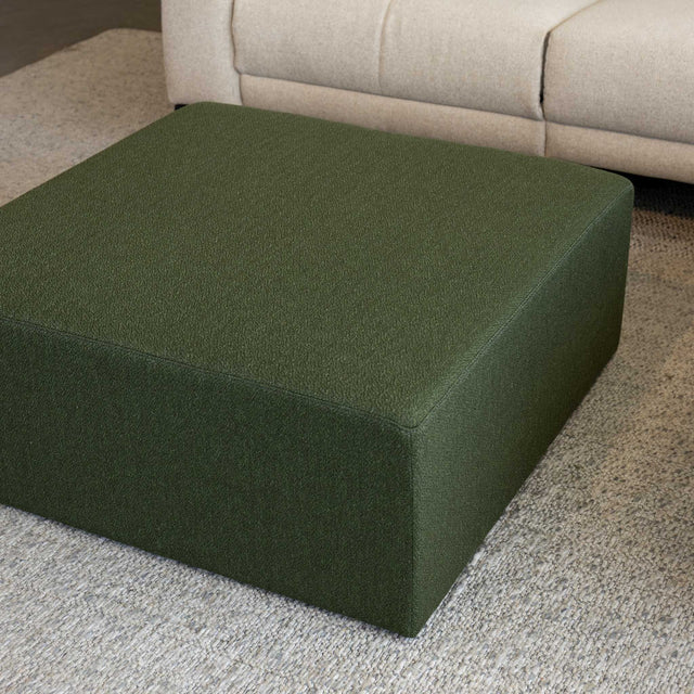 Ottoman carré vert en tissu, utilisé comme table basse devant un canapé d’angle moderne dans un salon contemporain.