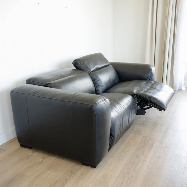 canape relax en cuir noir avec dossier inclinable et repose pieds ouvert sur sol en bois clair.
