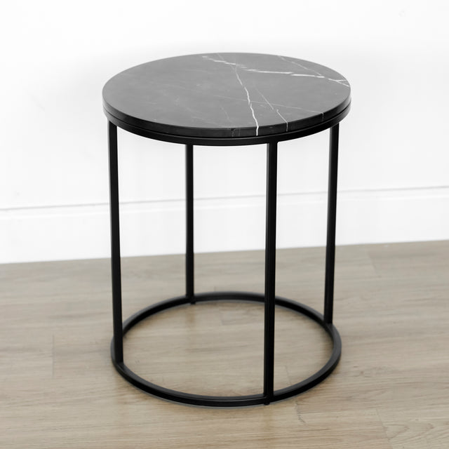 Table d’appoint ronde au design minimaliste, avec un plateau noir et une structure métallique fine de forme cylindrique, également noire.
