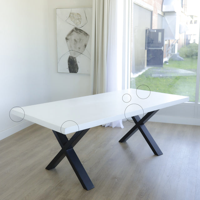 table moderne avec plateau blanc epais et pieds en metal noir en forme de x, sur parquet clair pres d’une grande fenetre.
