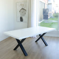 table moderne avec plateau blanc epais et pieds en metal noir en forme de x, sur parquet clair pres d’une grande fenetre.
