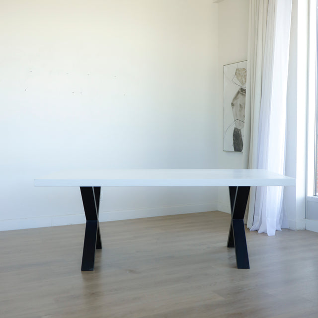 table rectangulaire blanche au design moderne, posée sur un sol en bois clair, avec des pieds croisés noirs et un fond de mur blanc éclairé par la lumière naturelle provenant d’une grande fenêtre.

