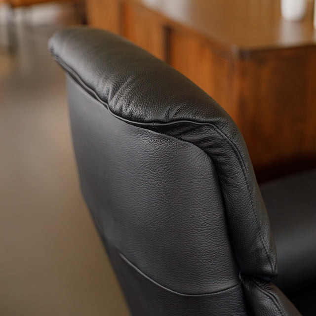 Fauteuil en cuir noir avec appui-tête et base bois pivotante, dans un intérieur chaleureux.