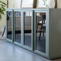 Buffet moderne vert sauge à portes vitrées fumées, décoré d’objets design dans un intérieur contemporain lumineux.