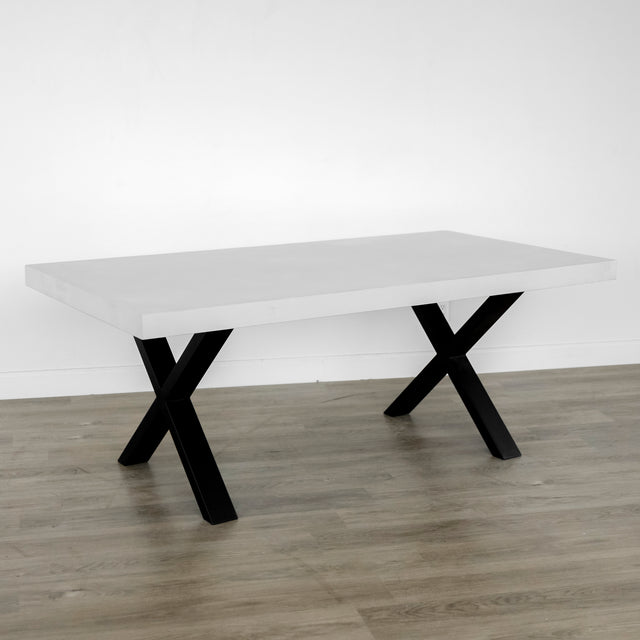 table rectangulaire moderne avec un plateau blanc mat épais et des pieds en métal noir en forme de X, posée sur un sol en bois clair devant un mur blanc.