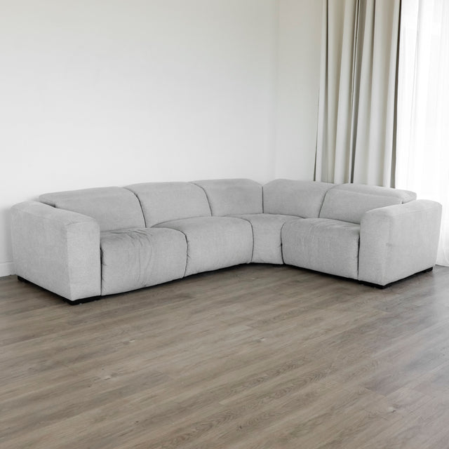canapé d’angle moderne en tissu gris clair à assise profonde, placé sur un sol en bois clair, devant un mur blanc avec de longs rideaux beiges.
