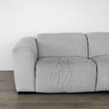canapé en tissu gris clair au design contemporain, avec accoudoirs larges et coussins d’assise moelleux, posé sur un sol en bois clair devant un mur blanc.
