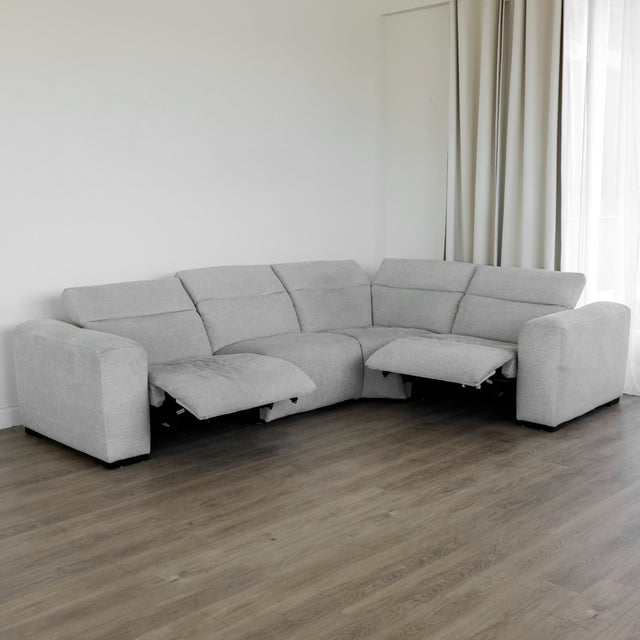 canapé d’angle en tissu gris clair avec deux assises inclinables dotées de repose-pieds relevés, design moderne et confort moelleux, placé sur un sol en bois clair près d’un mur blanc et de longs rideaux beiges.
