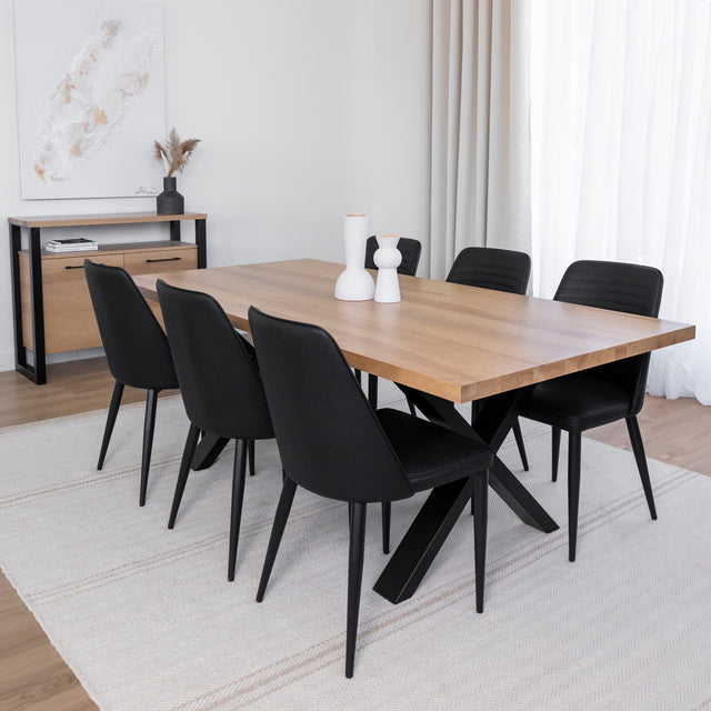 Table à manger en bois clair avec piètement en X métallique noir, entourée de six chaises noires dans une salle à manger moderne et épurée.