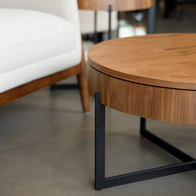 Table basse ronde en bois noyer avec rangement, près d’un fauteuil beige.