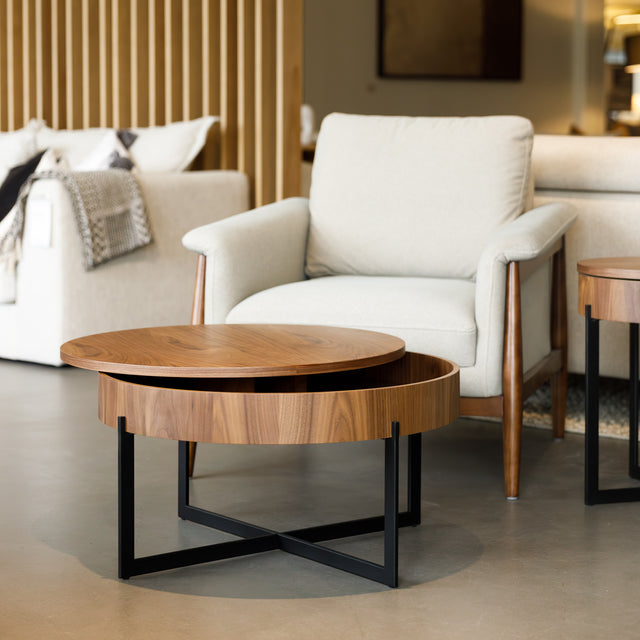 Table basse ronde en bois noyer avec rangement, près d’un fauteuil beige.