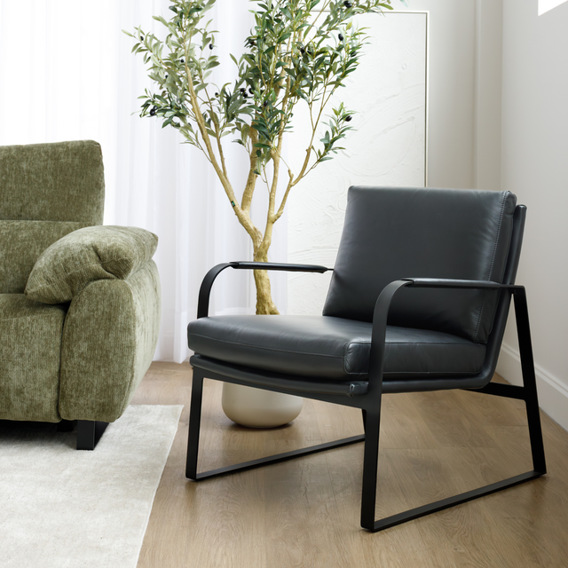 Fauteuil en cuir noir avec structure en métal, présenté dans un salon contemporain à côté d’un canapé en tissu vert.