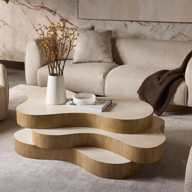 Table basse organique en bois clair à plateaux superposés, style sculptural, intégrée dans un salon avec canapé d’angle.