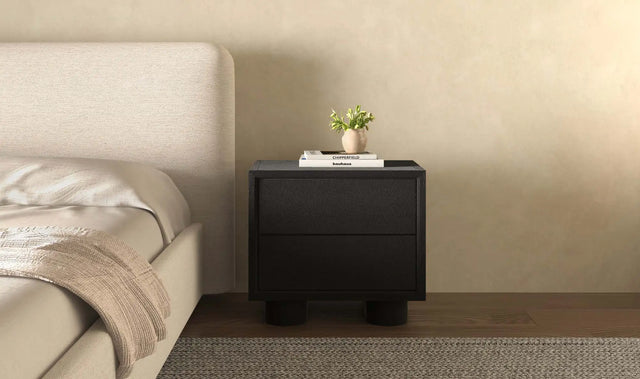 Table de chevet noire moderne avec deux tiroirs, posée à côté d’un lit beige dans une chambre minimaliste.