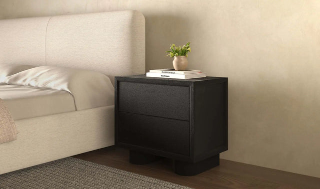 Table de chevet noire moderne avec deux tiroirs, posée à côté d’un lit beige dans une chambre minimaliste.