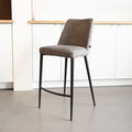 Tabouret de bar design avec assise en tissu brun/gris et pieds en metal noir, ideal pour cuisine moderne.